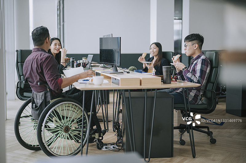 亚洲同事和印度同事在办公室工作站休息时，坐在轮椅上吃面包图片素材