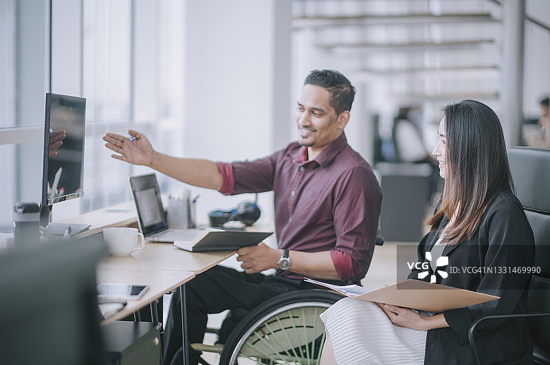 印度白领男员工坐在轮椅上，与中国女同事在创意办公室工作站愉快交谈图片素材