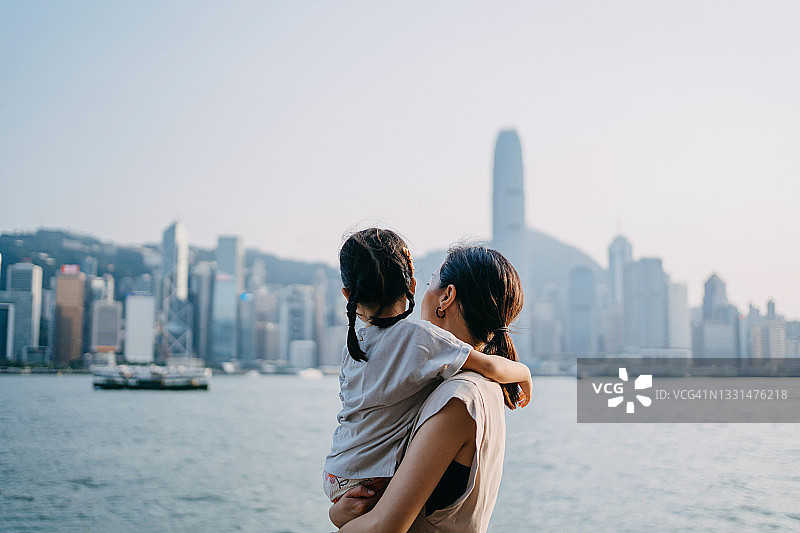 年轻的亚洲母亲在维多利亚港的滨海大道上拥抱可爱的女儿，欣赏壮观的城市天际线图片素材