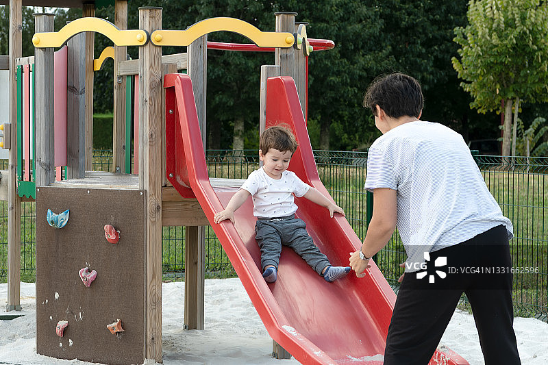 母亲和儿子在操场玩耍，孩子正在滑红色滑梯图片素材