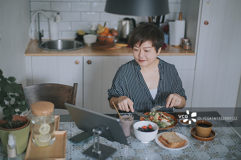 华人女性在餐桌旁享用健康沙拉早餐时，使用平板电脑收听音乐广播频道图片素材