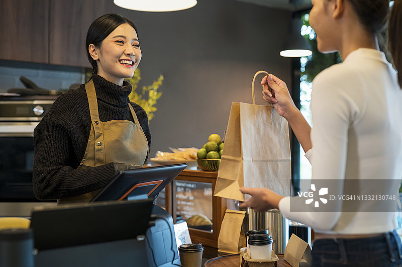 希望您喜欢您的咖啡。咖啡店女老板在收银台将外卖咖啡递给顾客，使用销售点系统图片素材