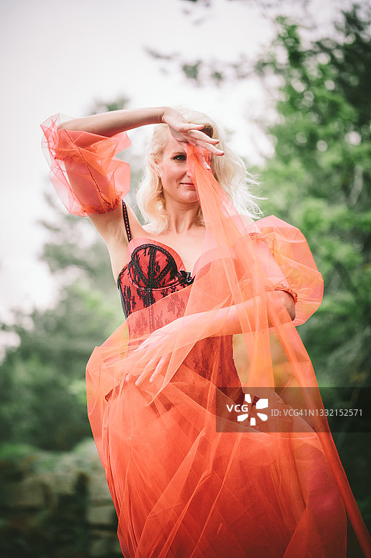 一位穿着舞厅红色连衣裙，裙摆飘逸的年轻金发女性，站在石头上，在树木中跳舞，为相机摆出舞蹈姿势。图片素材