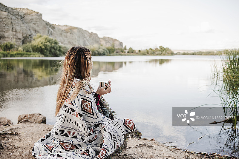 裹着毯子在湖边拿着咖啡杯的年轻金发女人图片素材