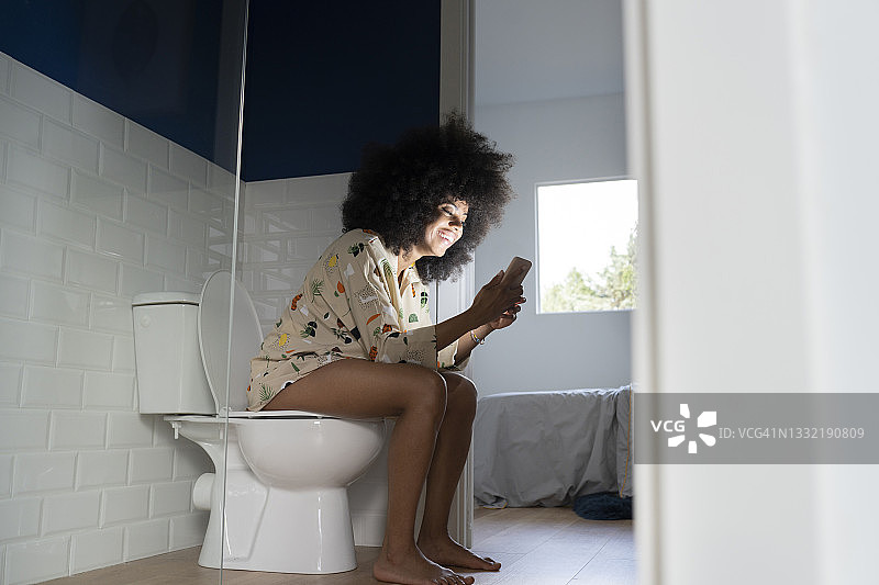 在家中坐在马桶上使用智能手机的微笑女人图片素材