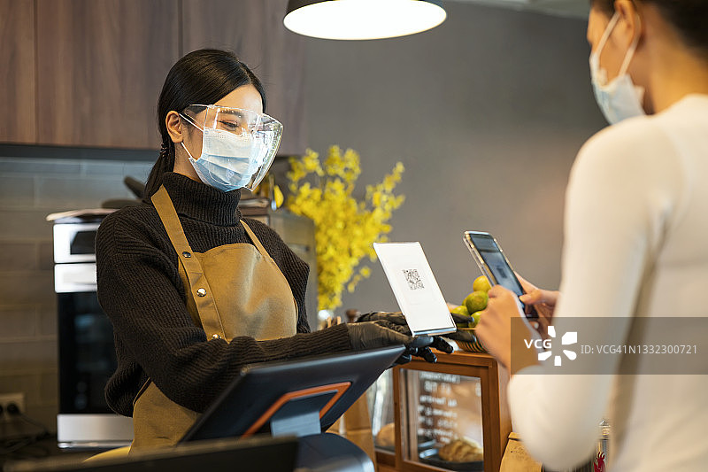 餐厅和咖啡馆安全提示：亚洲收银员展示条形码支付，顾客使用智能手机支付外卖咖啡。销售点系统、移动支付和数字钱包图片素材