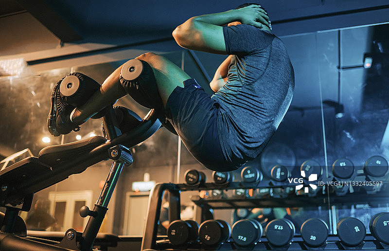 低角度视角：在夜晚的健身房里，印度男士在配重凳上做仰卧起坐图片素材