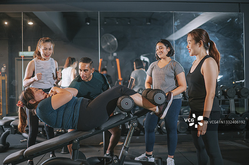 亚裔男健身教练指导年轻少女在健身房一起做仰卧起坐，还有其他女运动员图片素材