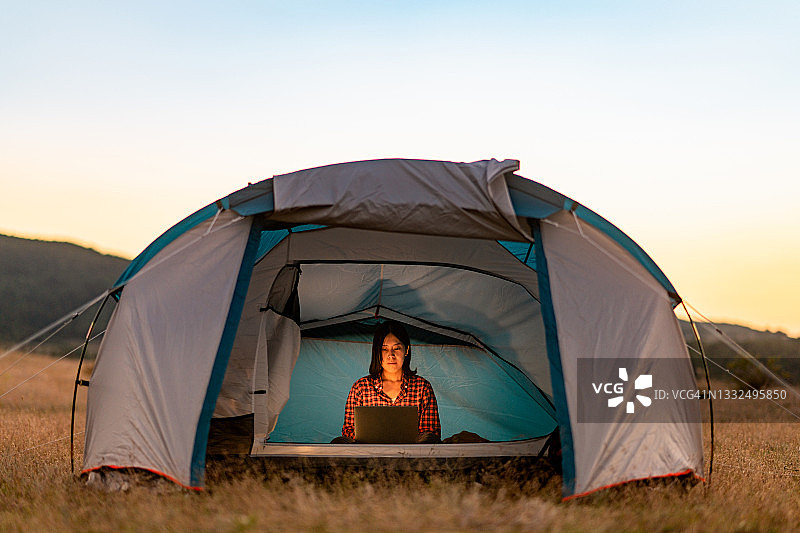 年轻女人在野外露营时在帐篷旁使用笔记本电脑图片素材