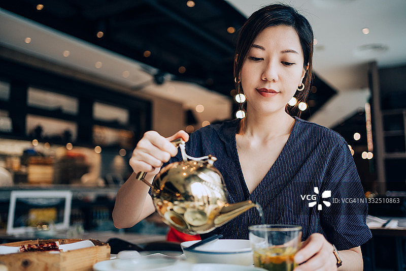 年轻的亚洲女性在中餐馆享用新鲜的传统点心。她正在餐桌上往杯子里倒中国茶。传统中国文化，饮茶，外出就餐生活方式图片素材