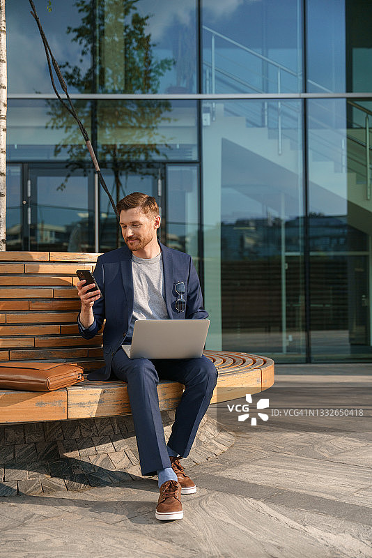 身穿蓝色西装和灰色T恤的有魅力的年轻商人在商务中心附近的圆形长椅上，膝盖上放着笔记本电脑，手里拿着智能手机图片素材