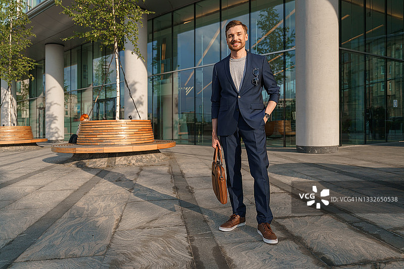 一位穿着蓝色商务套装的年轻有魅力的商人在现代商务中心背景下手提皮质笔记本电脑包图片素材
