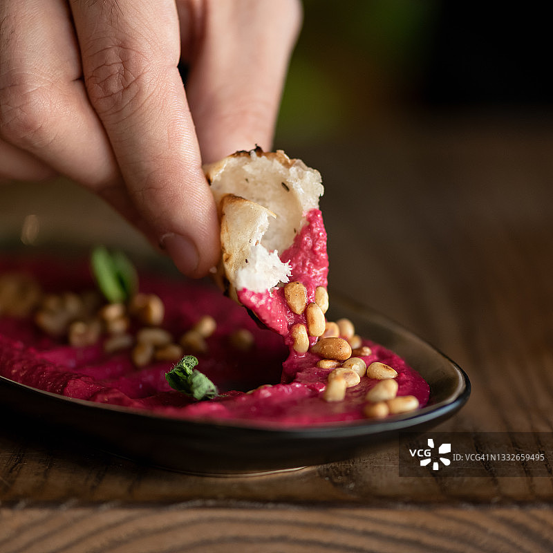 男人的手将皮塔饼蘸入装有红色鹰嘴豆泥或蘸酱的碗中。甜菜根泥配花生，特写镜头。柔焦。复制空间图片素材