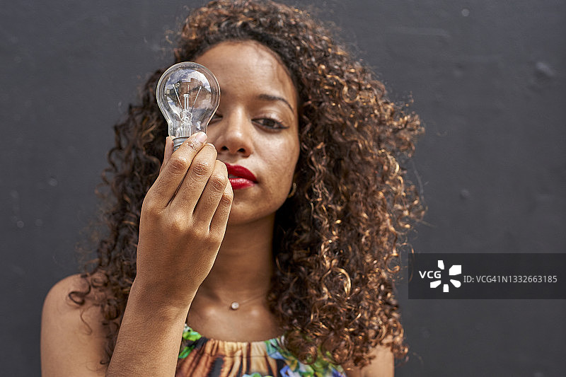 年轻的非洲裔卷发女人穿着绿色露脐上衣，在黑墙前拿着灯泡图片素材
