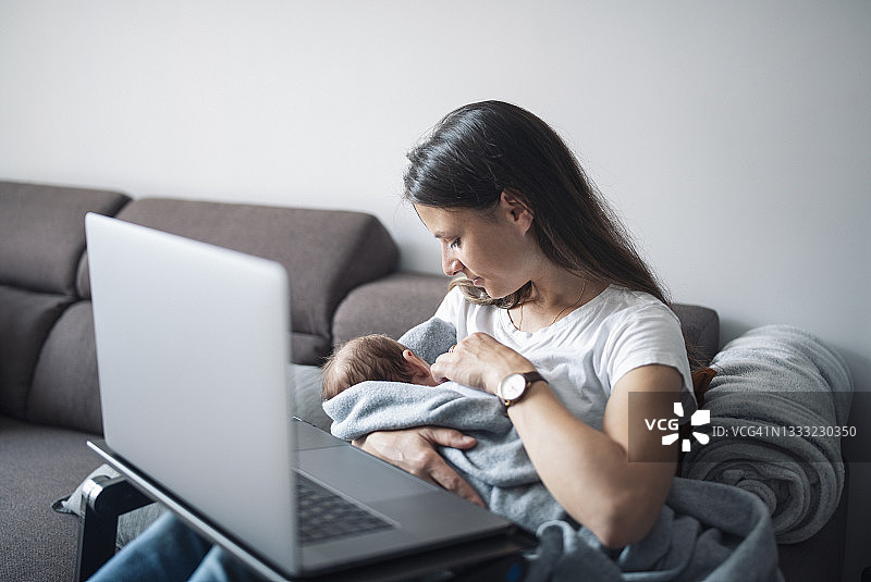 年轻的母亲在客厅里用笔记本电脑给新生儿喂奶图片素材