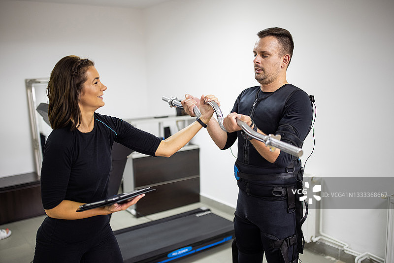 健身教练向穿着肌肉电刺激服的男子讲解，同时该男子在健身房用杠铃锻炼图片素材