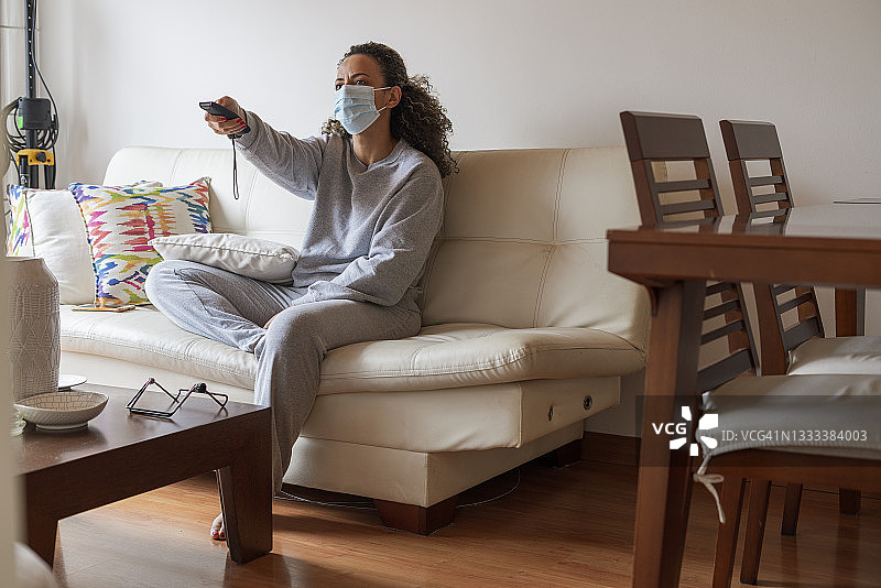 女人戴着口罩坐在客厅的沙发上，用遥控器看电视图片素材
