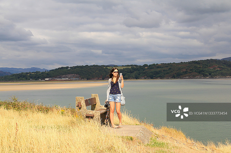 海边自拍：年轻女子在阳光明媚的日子站在悬崖顶上用手机自拍，发给朋友图片素材