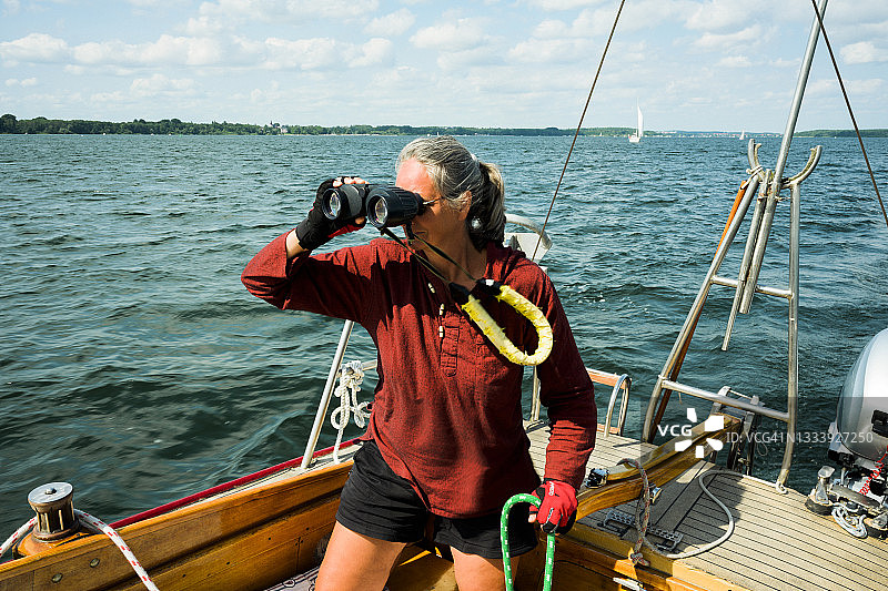 活跃的中年妇女船长在德国驾驶帆船，用双筒望远镜观察图片素材