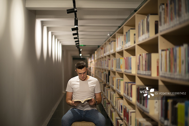 戴眼镜的年轻男学生在图书馆走廊里看书图片素材