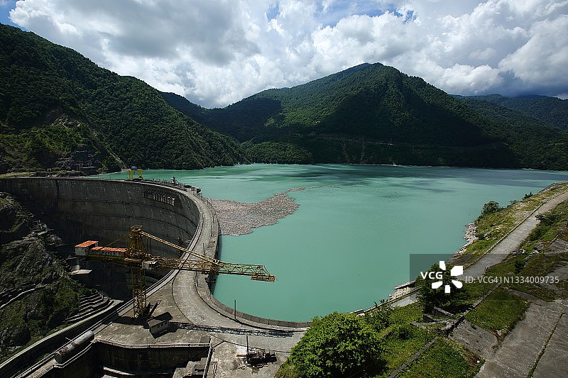 多云天空下碧绿色水坝湖的景色，背景是绿色森林山脉图片素材