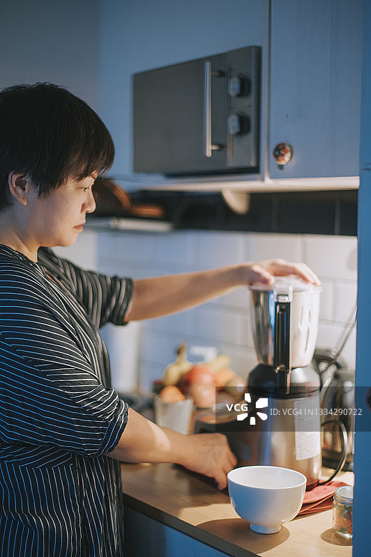 高角度侧视：亚洲中年妇女在厨房用搅拌机准备健康冰沙图片素材