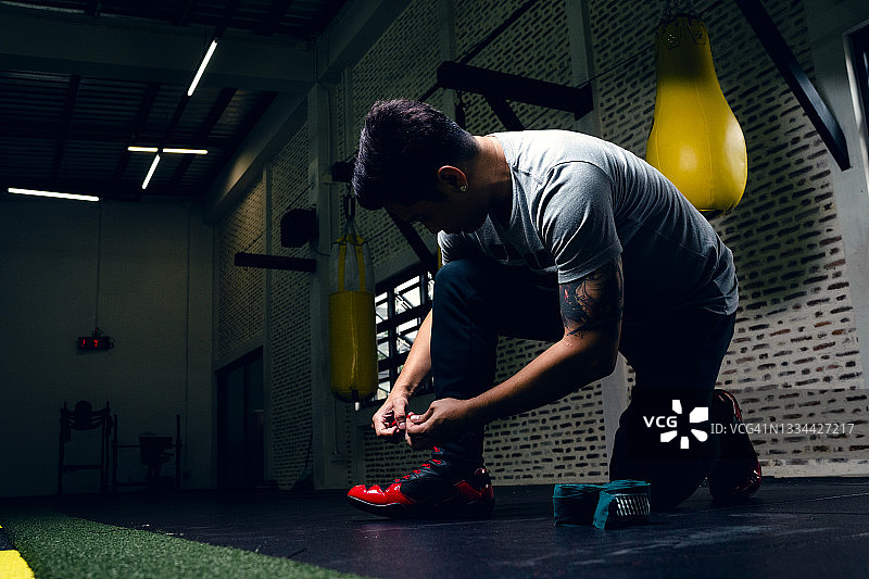年轻男子系紧鞋带，准备开始拳击训练，背景是拳击沙袋图片素材