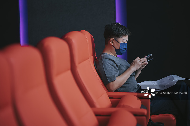 影院重新开放时，戴口罩的亚洲中年华人女性在电影院使用智能手机的侧视图图片素材