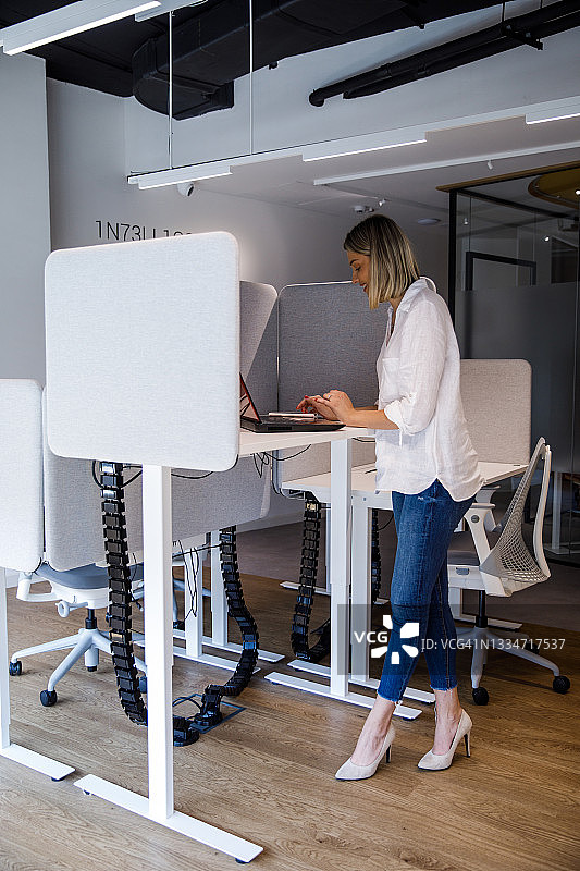 在办公室隔间使用站立式办公桌的女商人，在笔记本电脑上录入数据图片素材