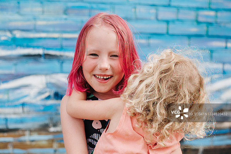 年龄不同的姐妹拥抱、玩耍和大笑。一个是有粉色头发的学龄期姐姐，另一个是穿着粉色外套的幼儿园妹妹。图片素材