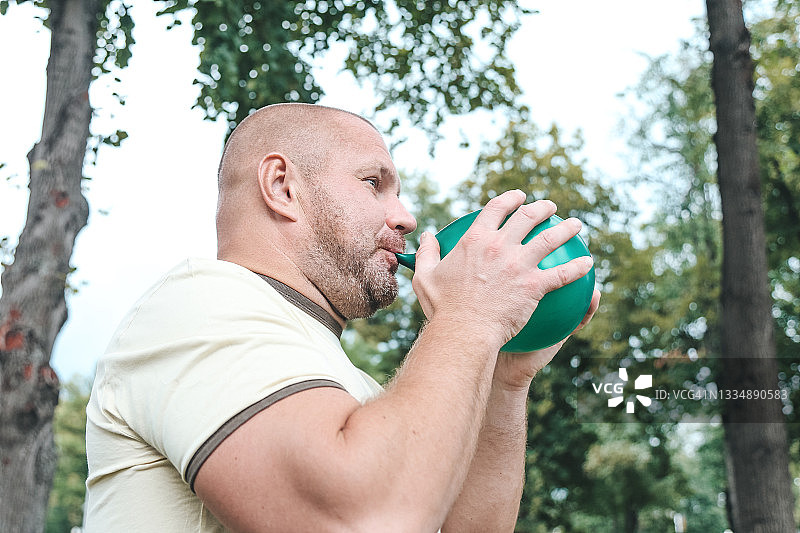 在绿树背景下吹气球的成年秃顶男人图片素材