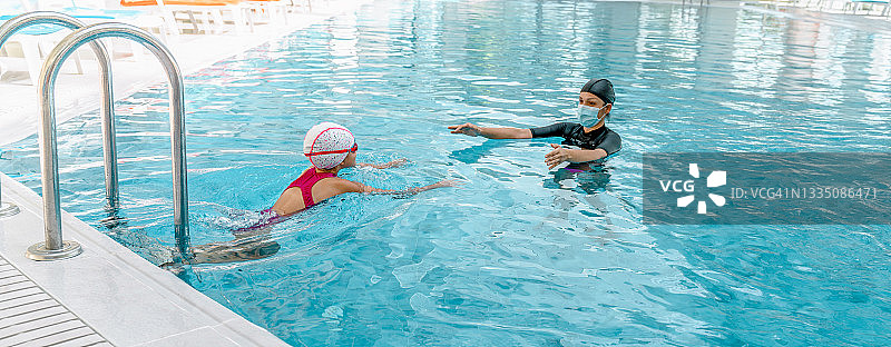 女教练戴着面部医用防护口罩在室内游泳池教孩子游泳图片素材