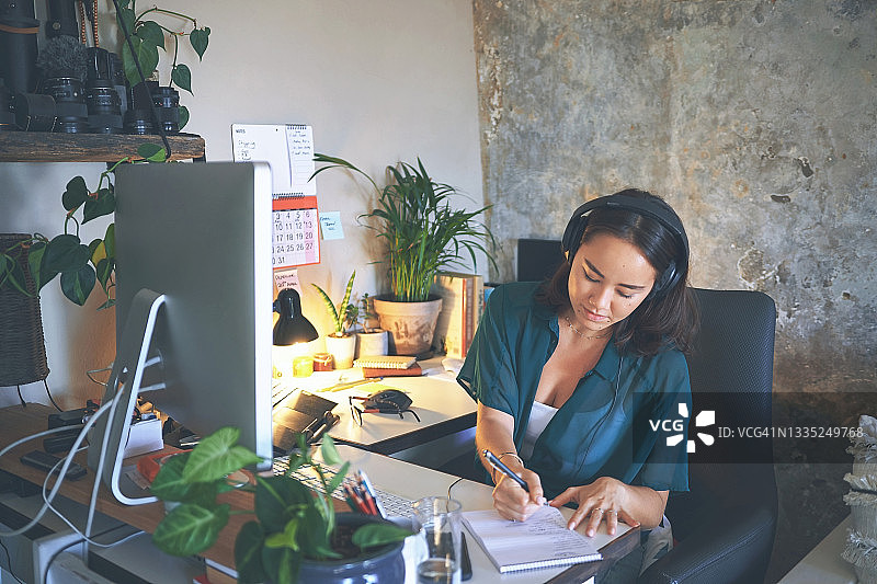 年轻女子在家庭办公室戴着耳机写笔记图片素材