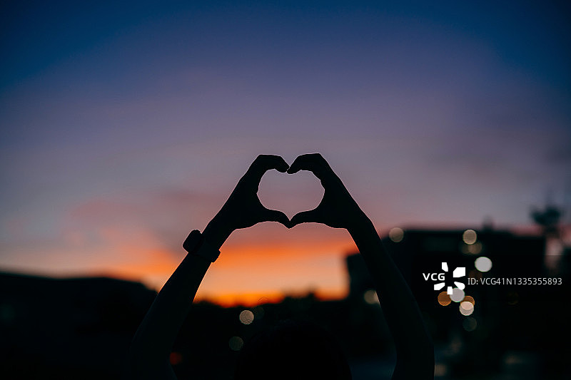 黄昏时分，女人举起手在引人注目的天空上做出心形的特写。爱、关怀、希望和积极的氛围概念图片素材