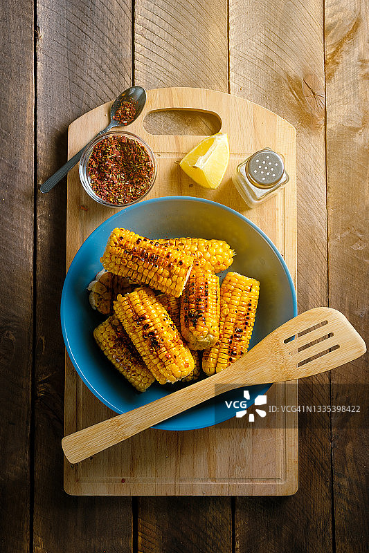 烤制的黄色玉米棒，撒上香料，放在明亮的陶瓷盘中，木桌背景。自制食物烹饪概念。图片素材