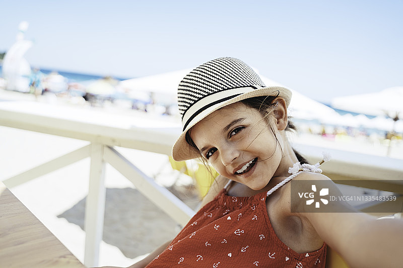 戴着帽子的可爱女孩坐在沙滩遮阳伞下，在暑假自拍。穿着休闲的孩子在热带天堂海滩放松身心，享受假期。图片素材