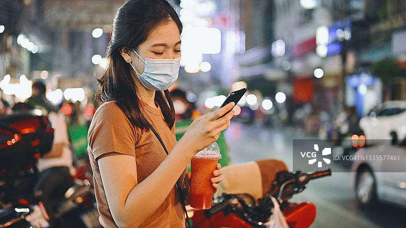 年轻漂亮的亚洲女性在曼谷耀华力路使用手机APP等待出租车/公共汽车，她戴着口罩以保护自己免受新冠病毒感染，背景是曼谷和嘟嘟出租车图片素材