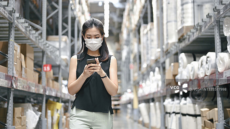 亚洲女商人使用手机应用程序比较现代贸易价格，检查大型仓库中的预订单。在线卖家进出口图片素材
