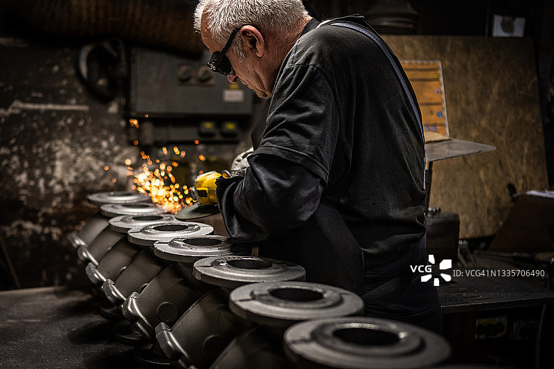年长的男人在黑暗和多尘的工作场所工作，打磨铸件图片素材