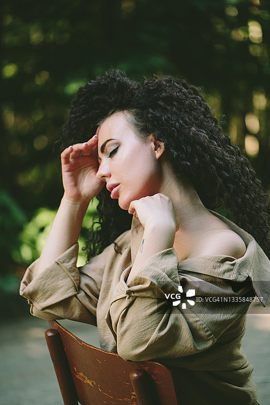 一位留着爆炸头的年轻白人棕发女孩身穿米色休闲西装，坐在马路中央的复古椅子上，地平线两侧是绿树。侧视图。图片素材