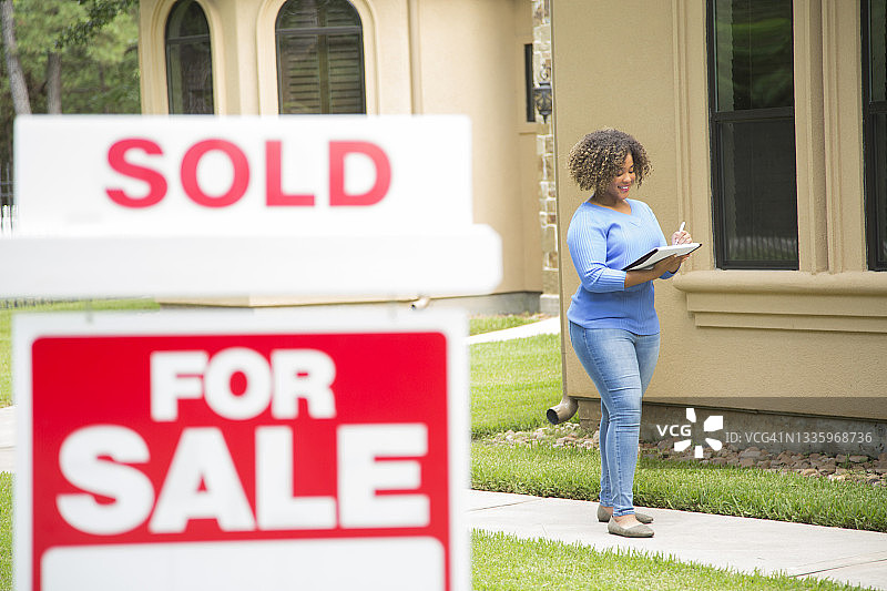 年轻的房地产经纪人站在自家院子里的待售标志旁边，拿着文件夹图片素材