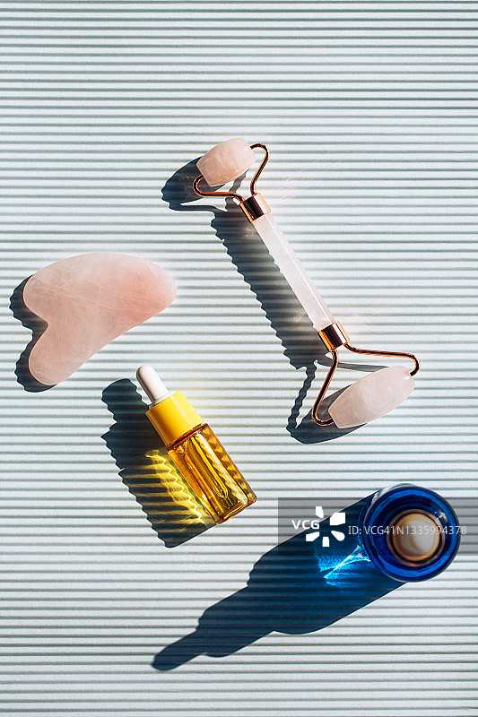 两瓶玻璃瓶、玫瑰石英滚轮和刮痧板，置于条纹灰色背景上。用于皮肤护理的面部精华液或保湿霜。阳光在瓶子上形成阴影和反射。扁平风格。图案图片素材