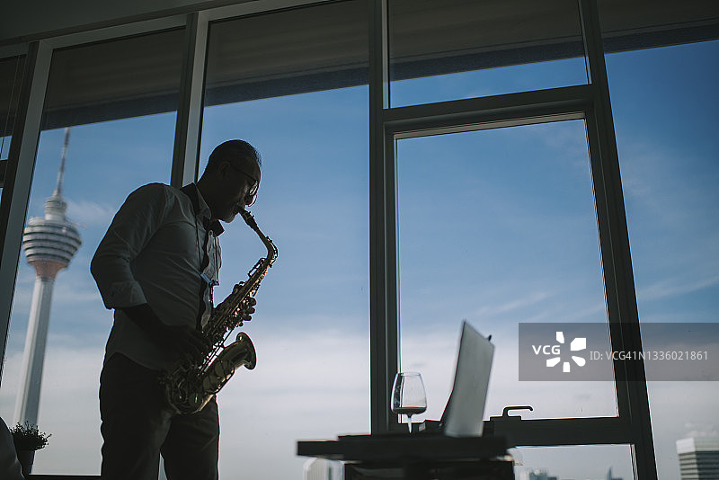 在剪影中，亚裔活跃老年男人艺术家在客厅里用笔记本电脑演奏萨克斯并展示给他的学生看图片素材