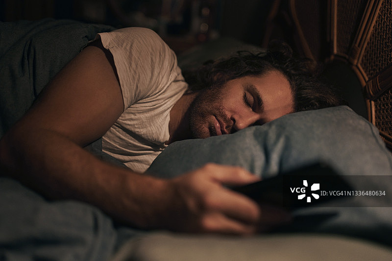 在家中床上，手持手机睡觉的年轻男子特写图片素材
