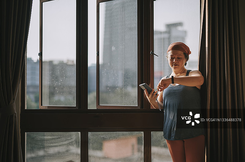 亚裔中年女性戴头巾，在阳光明媚的窗边使用智能手机记录健康状态图片素材