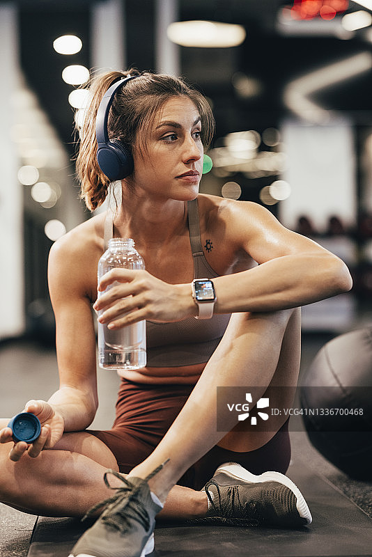 美丽的白人女运动员在健身房的垫子上听音乐，手里拿着一瓶水图片素材
