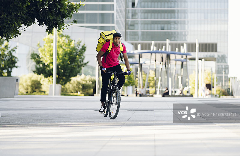笑容满面的送货员背着黄色背包在城市里骑自行车图片素材