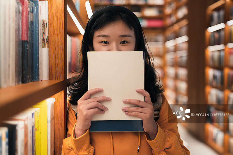 图书馆：亚洲女子在书架旁用书遮面微笑图片素材