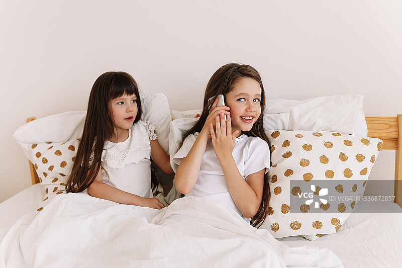 两个穿着睡衣的小女孩在舒适的卧室里看手机和笔记本电脑自拍，与父母沟通，在家上网课。图片素材