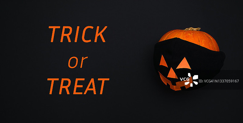 黑色背景上的南瓜，戴着黑色防护面罩，面罩上有橙色纸做的眼睛、嘴巴和鼻子，黑色背景上写着“不给糖就捣蛋！”万圣节和新冠肺炎概念图片素材